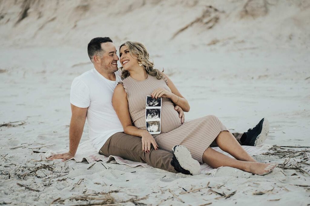 Couple sitting in sand on beach holding sonogram for maternity session