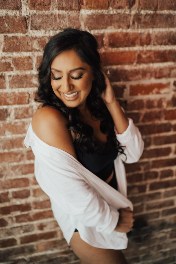 Boudoir photoshoot with woman standing against brick wall with hand in hair.